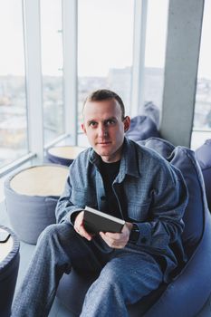 A young male student in a casual suit resting in a comfortable pouf while studying makes notes in a notebook, an attractive freelancer in a suit works remotely