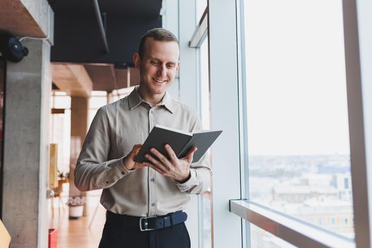 A young businessman stands at a panoramic window in a skyscraper and makes notes in his notebook with a pen. A man in a shirt and trousers of European appearance