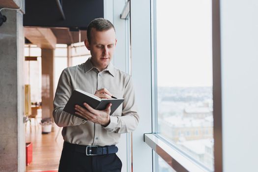 A young businessman stands at a panoramic window in a skyscraper and makes notes in his notebook with a pen. A man in a shirt and trousers of European appearance