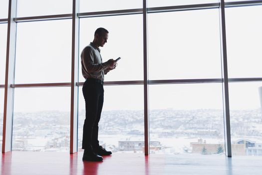 A young executive is holding a phone while standing in an office interior and looking out a large window overlooking the city. Male manager with a phone is thinking about a new business project