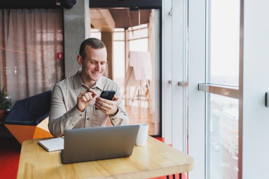 Successful young man is sitting at a table in a cafe-restaurant indoors, working and studying on a laptop computer,holding a mobile phone with a blank screen. Freelancer mobile office business concept