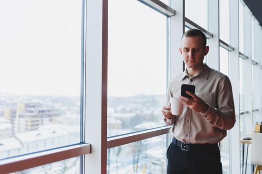 Male brokers with phone looking towards window with coffee in hand in modern workspace
