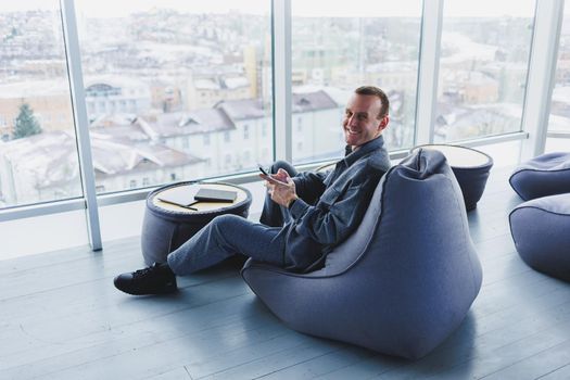 Break. Smiling young man relaxing on bean bag chair while sitting at table and relaxing using laptop, happy businessman leaning back, enjoying work, feeling satisfied