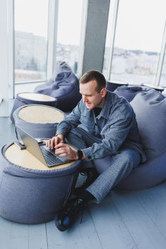 Break. Smiling young man relaxing on bean bag chair while sitting at table and relaxing using laptop, happy businessman leaning back, enjoying work, feeling satisfied