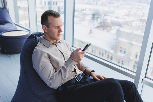 Successful male freelancer sitting in modern workspace and checking notifications on mobile phone in casual clothes. Remote work.