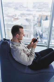 Successful male businessman sitting in a modern workspace and checking notifications on his mobile phone, he is wearing a shirt and trousers. Remote work.