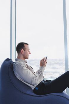 Successful male businessman sitting in a modern workspace and checking notifications on his mobile phone, he is wearing a shirt and trousers. Remote work.