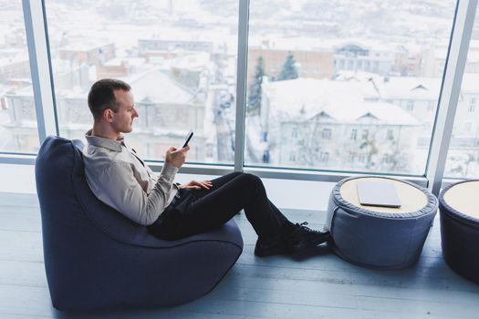 Successful male freelancer sitting in modern workspace and checking notifications on mobile phone in casual clothes. Remote work.