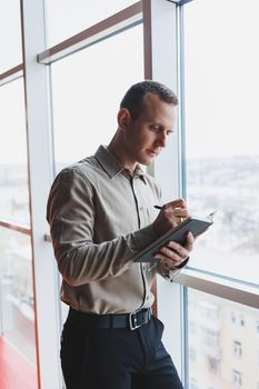 A young businessman stands at a panoramic window in a skyscraper and makes notes in his notebook with a pen. A man in a shirt and trousers of European appearance