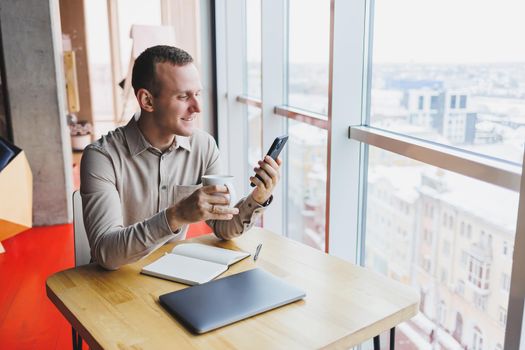 Successful young man is sitting at a table in a cafe-restaurant indoors, working and studying on a laptop computer,holding a mobile phone with a blank screen. Freelancer mobile office business concept