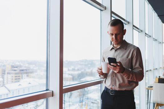 Male brokers with phone looking towards window with coffee in hand in modern workspace