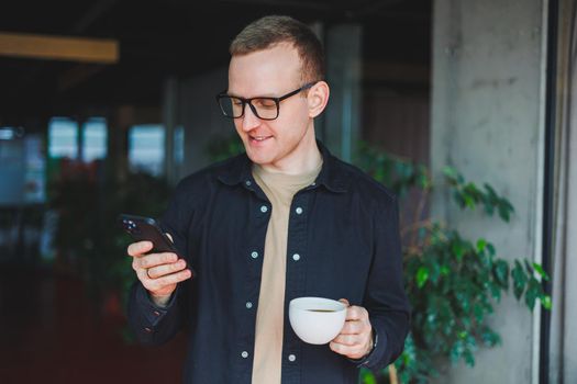 Portrait of a happy successful man in optical glasses holding a modern mobile phone and coffee in his hands, a cheerful blogger with a digital smartphone posing in a cafe