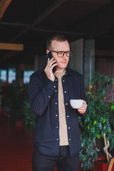 Portrait of a happy successful man in optical glasses holding a modern mobile phone and coffee in his hands, a cheerful blogger with a digital smartphone posing in a cafe