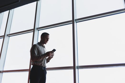 A young executive is holding a phone while standing in an office interior and looking out a large window overlooking the city. Male manager with a phone is thinking about a new business project