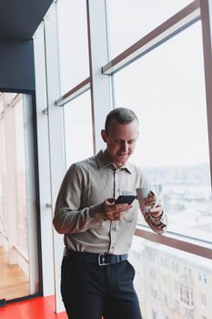 Male brokers with phone looking towards window with coffee in hand in modern workspace