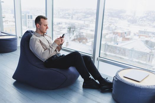 Successful male freelancer sitting in modern workspace and checking notifications on mobile phone in casual clothes. Remote work.