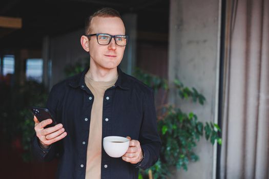 Portrait of a happy successful man in optical glasses holding a modern mobile phone and coffee in his hands, a cheerful blogger with a digital smartphone posing in a cafe