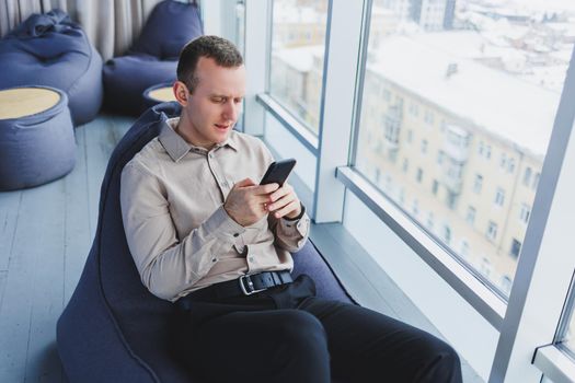 Successful male freelancer sitting in modern workspace and checking notifications on mobile phone in casual clothes. Remote work.