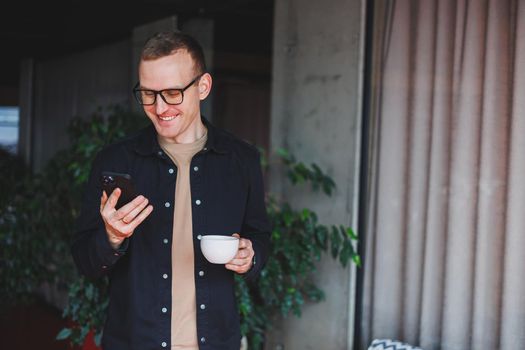 Portrait of a happy successful man in optical glasses holding a modern mobile phone and coffee in his hands, a cheerful blogger with a digital smartphone posing in a cafe