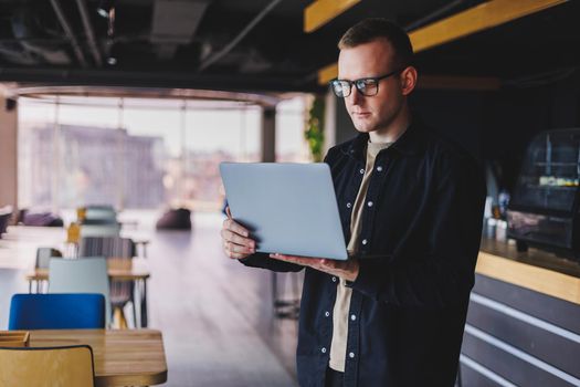 Male entrepreneur in black shirt and glasses standing in office with laptop in hand, successful corporate boss feeling good from rich lifestyle