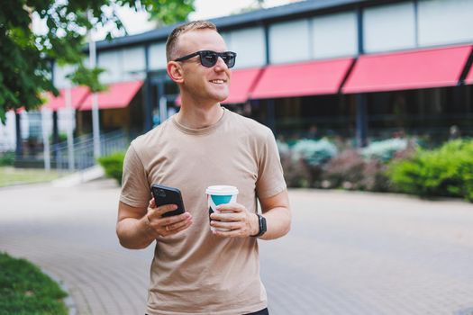 A bearded, smiling, stylish man in white shirt and sunglasses standing on the streets of the city and calling on mobile phone.