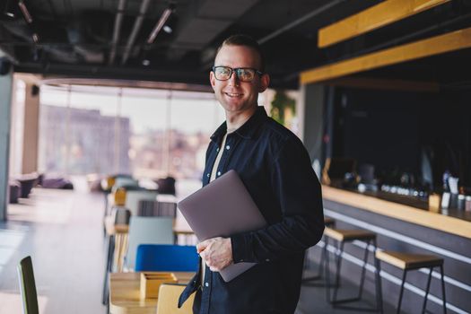 Male entrepreneur in black shirt and glasses standing in office with laptop in hand, successful corporate boss feeling good from rich lifestyle