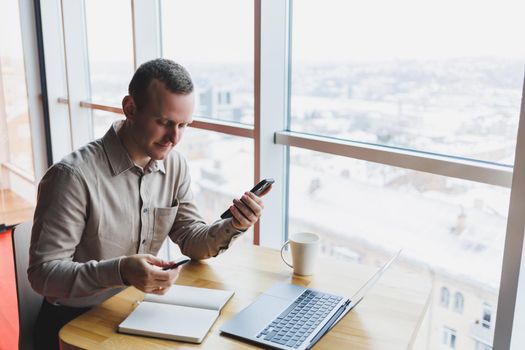 Successful young man is sitting at a table in a cafe-restaurant indoors, working and studying on a laptop computer,holding a mobile phone with a blank screen. Freelancer mobile office business concept