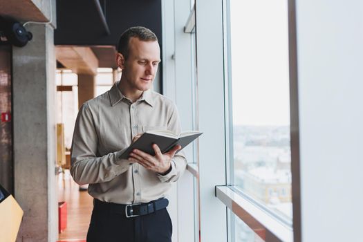 A young businessman stands at a panoramic window in a skyscraper and makes notes in his notebook with a pen. A man in a shirt and trousers of European appearance