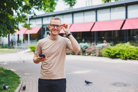 Portrait of handsome mature businessman holding messenger bag, wearing stylish eyeglasses looking at camera. Smiling middle aged man using smartphone, standing on the street. Mobile banking concept