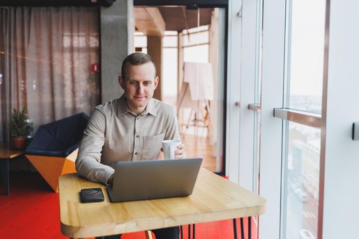 A young smart man is sitting in the office with a cup of coffee and is working on a project related to public cyber technology. Businessmen with laptop trying to meet deadlines in external marketing.