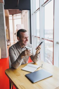 Successful young man is sitting at a table in a cafe-restaurant indoors, working and studying on a laptop computer,holding a mobile phone with a blank screen. Freelancer mobile office business concept