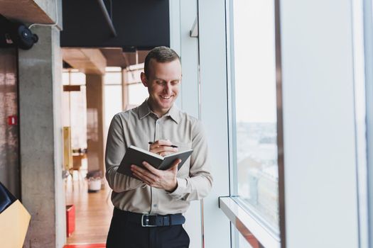 A young businessman stands at a panoramic window in a skyscraper and makes notes in his notebook with a pen. A man in a shirt and trousers of European appearance