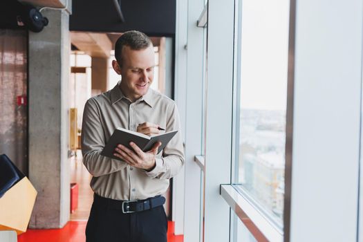 A young businessman stands at a panoramic window in a skyscraper and makes notes in his notebook with a pen. A man in a shirt and trousers of European appearance