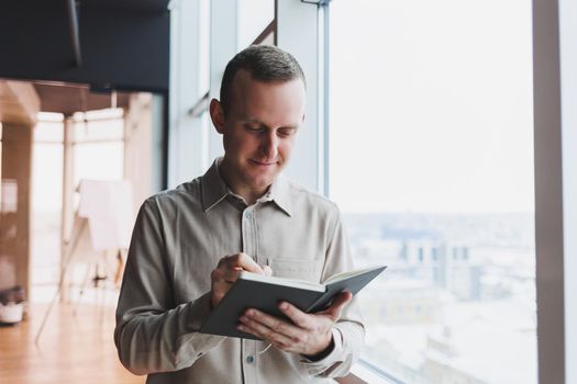 A young businessman stands at a panoramic window in a skyscraper and makes notes in his notebook with a pen. A man in a shirt and trousers of European appearance