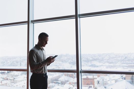 A young executive is holding a phone while standing in an office interior and looking out a large window overlooking the city. Male manager with a phone is thinking about a new business project