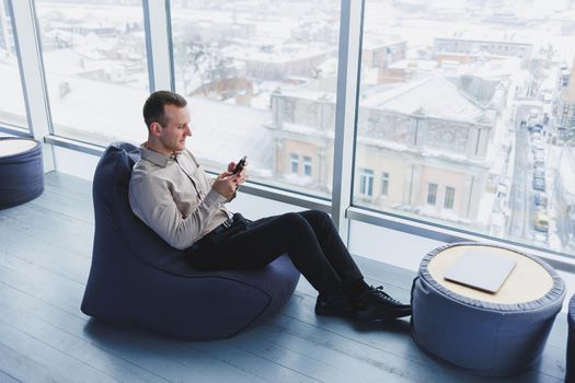 Successful male freelancer sitting in modern workspace and checking notifications on mobile phone in casual clothes. Remote work.