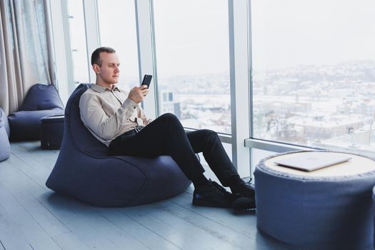 Successful male freelancer sitting in modern workspace and checking notifications on mobile phone in casual clothes. Remote work.