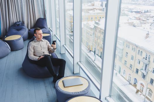 Successful male businessman sitting in a modern workspace and checking notifications on his mobile phone, he is wearing a shirt and trousers. Remote work.