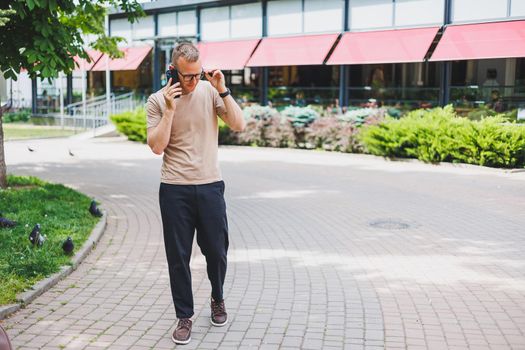 Portrait of handsome mature businessman holding messenger bag, wearing stylish eyeglasses looking at camera. Smiling middle aged man using smartphone, standing on the street. Mobile banking concept