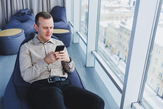 Successful male businessman sitting in a modern workspace and checking notifications on his mobile phone, he is wearing a shirt and trousers. Remote work.