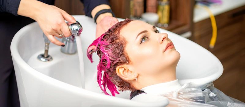 Washing dyed female hair. A young caucasian woman having her hair washed in a beauty salon. Professional hairdresser washes pink color paint off of a customer's hair