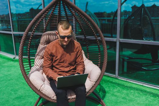 A businessman in formal wear sits on an armchair and looks at a laptop connected to a 4g wireless network on the terrace of a modern office while working remotely