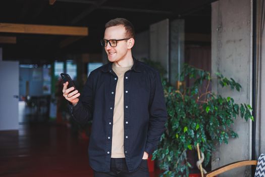 Half-length portrait of happy man in optical glasses holding modern mobile phone and smiling at camera, cheerful blogger with digital smartphone posing in coworking space