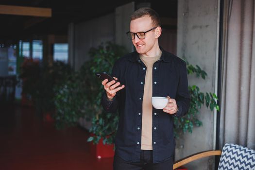 Portrait of a happy successful man in optical glasses holding a modern mobile phone and coffee in his hands, a cheerful blogger with a digital smartphone posing in a cafe