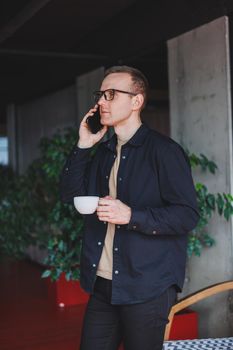 Portrait of a happy successful man in optical glasses holding a modern mobile phone and coffee in his hands, a cheerful blogger with a digital smartphone posing in a cafe