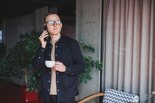 Portrait of a happy successful man in optical glasses holding a modern mobile phone and coffee in his hands, a cheerful blogger with a digital smartphone posing in a cafe