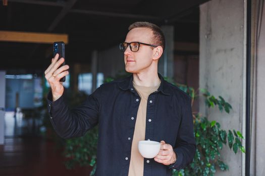 Portrait of a happy successful man in optical glasses holding a modern mobile phone and coffee in his hands, a cheerful blogger with a digital smartphone posing in a cafe