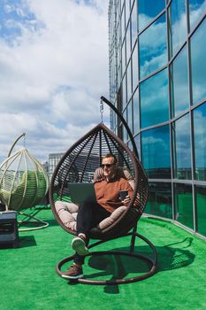 A businessman in formal wear sits on an armchair and looks at a laptop connected to a 4g wireless network on the terrace of a modern office while working remotely