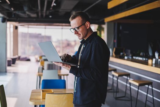 Male entrepreneur in black shirt and glasses standing in office with laptop in hand, successful corporate boss feeling good from rich lifestyle