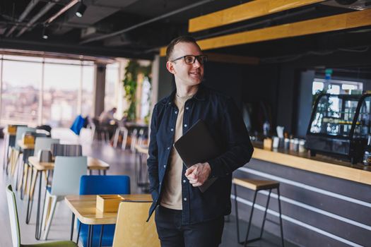 Male entrepreneur in black shirt and glasses standing in office with laptop in hand, successful corporate boss feeling good from rich lifestyle